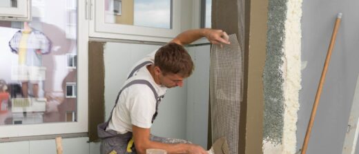 Un artisan applique un mur, des outils autour. L'accueil est visible par la fenêtre.